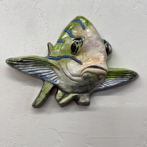 Op de afbeelding: Een keramische vissenmuurhanger met een gedetailleerd ontwerp. De vis heeft een groen lichaam met blauwe strepen, een wit en perzikkleurig gezicht en grote zwarte ogen. De vinnen zijn groen en wit, en de vis heeft een enigszins humeurige uitdrukking.