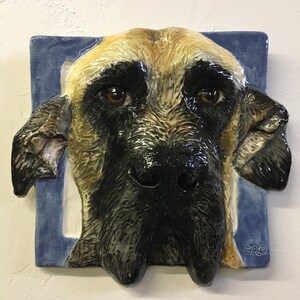 Può includere: Una scultura da parete in ceramica di una testa di cane. Il cane ha un pelo corto, marrone e nero, con un grande naso nero. La scultura è montata su uno sfondo blu.