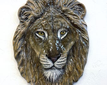 May include: A detailed lion head sculpture in shades of brown and tan, featuring a textured mane and yellow eyes. The sculpture is mounted on a white background, highlighting the intricate details. The lion's muzzle is white, adding contrast to the overall design.