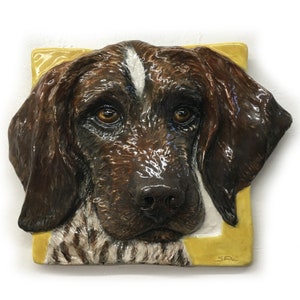 Könnte beinhalten: Keramikfliese mit einer detaillierten, dreidimensionalen Darstellung eines Hundekopfes. Der Hund hat braunes und weißes Fell, einen weißen Streifen auf der Stirn und ausdrucksstarke braune Augen. Die Fliese ist vor einem gelben Hintergrund angeordnet.