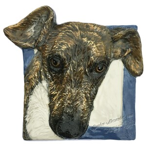 Può includere: Una piastrella in ceramica con la testa di un cane marrone e bianco che sporge da una cornice blu. Il cane ha un'espressione seria e i suoi occhi guardano direttamente lo spettatore. La piastrella è firmata "SRA" e ha il testo "Sandra Alexander.com Copyright" nell'angolo in basso a destra.