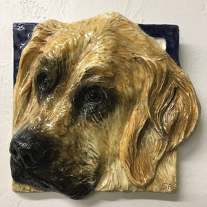 Può includere: Scultura a rilievo in ceramica raffigurante la testa di un cane, con una dettagliata rappresentazione del muso del cane. La scultura ha una forma quadrata con un bordo blu scuro. Il pelo del cane è reso in tonalità beige e marrone.