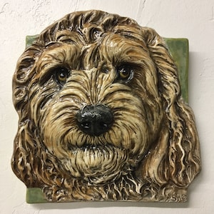 May include: A ceramic wall hanging featuring a detailed, three-dimensional depiction of a dog's face. The dog has brown fur, a black nose, and expressive brown eyes. The artwork is mounted on a square, with a green border at the bottom.