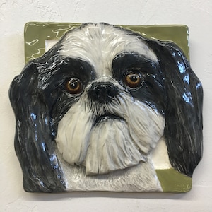 Op de afbeelding: Een keramische wandplaquette met een zwart-witte Shih Tzu hond met bruine ogen. Het gezicht van de hond is omlijst door een groene rand.