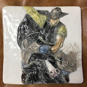 Könnte beinhalten: Eine Keramikfliese, die einen Cowboy auf einem sich aufbäumenden Bronco zeigt. Der Cowboy trägt eine schwarze Weste, ein gelbes Hemd und einen braunen Cowboyhut. Der Bronco ist schwarz-weiß mit einem braunen Sattel. Die Fliese ist weiß mit einer strukturierten Oberfläche.