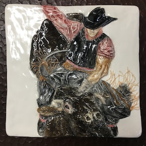 Könnte beinhalten: Keramikreliefskulptur einer Rodeo-Szene. Ein Cowboy mit schwarzem Hut und Weste reitet einen Bullen. Die Skulptur befindet sich auf einer weißen quadratischen Fliese mit braunem Rand. Der Cowboy trägt ein rotes Hemd.