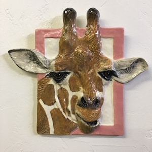 Könnte beinhalten: Eine Keramik-Giraffe-Kopfskulptur mit einem rosafarbenen Rahmen. Die Giraffe hat braune und weiße Markierungen und einen freundlichen Ausdruck.