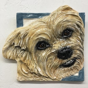 Op de afbeelding: Een keramische sculptuur van een hondengezicht, met gedetailleerde vachttextuur in tinten van bruin en crème. De ogen van de hond zijn bruin en de neus is zwart. De sculptuur is geplaatst in een blauw vierkant frame.