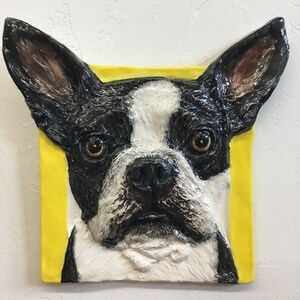 Peut inclure: Un gros plan d'une sculpture en céramique représentant le visage d'un Boston Terrier. Le chien a un pelage noir et blanc et regarde directement le spectateur. La sculpture est placée sur un fond jaune.