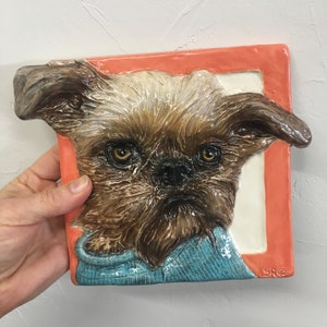 May include: A ceramic sculpture of a brown and white dog wearing a blue sweater. The sculpture is framed in a red and white frame with the signature "SRO" in the bottom right corner.