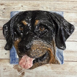 Può includere: Una scultura da parete in ceramica della testa di un Rottweiler con un manto nero e marrone, occhi marroni e una lingua rosa che sporge. La scultura è su uno sfondo blu con la firma "SRO" nell'angolo in basso a sinistra.