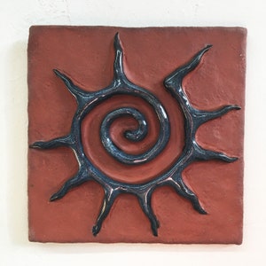 May include: A terracotta tile featuring a blue and black sun design. The sun has a spiral in the centre and rays extending outwards.