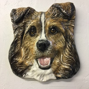 Può includere: Una scultura da parete in ceramica di una testa di cane. Il cane ha il pelo marrone, nero e bianco e sorride con la lingua di fuori.