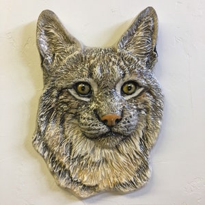 Könnte beinhalten: Eine Keramik-Wandskulptur eines Luchs-Kopfes. Die Skulptur ist in Braun-, Weiß- und Schwarztönen bemalt, mit realistischen Details. Der Luchs hat große, gelbe Augen und einen flauschigen, weißen Bart.