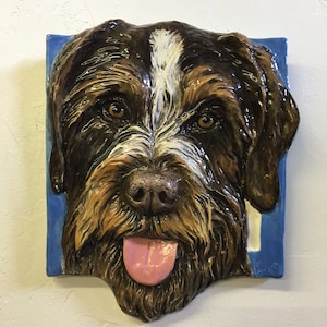 Può includere: Una scultura in ceramica di una testa di cane marrone e bianco con la lingua rosa che sporge. Il cane ha un motivo di pelliccia marrone e bianco e un naso nero. La scultura è su uno sfondo blu.