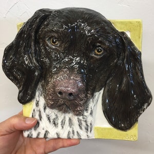 Può includere: Una scultura in ceramica di una testa di Bracco tedesco a pelo corto, di colore marrone e bianco. La scultura è montata su uno sfondo giallo con la firma dell'artista "SAO" in basso a destra.