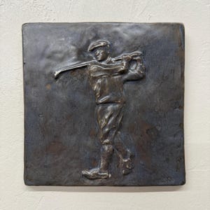 Op de afbeelding: Een bronzen reliëfsculptuur van een golfer in volle swing, geplaatst tegen een vierkante, getextureerde achtergrond. De figuur draagt een pet en golfkleding, de club is geheven. Het kunstwerk is op een lichtgekleurde muur gemonteerd.