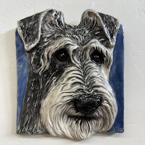 Op de afbeelding: Een keramische wanddecoratie met een gedetailleerd portret van een Schnauzer-hond. De sculptuur heeft een getextureerd, driedimensionaal uiterlijk met zwart, grijs en wit haar, bruine ogen en een zwarte neus. De hond staat tegen een blauwe achtergrond.