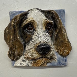 Può includere: Una piastrella in ceramica con una rappresentazione dettagliata e tridimensionale del volto di un cane. Il cane ha pelo marrone e bianco, un naso nero e occhi marroni espressivi. La piastrella è ambientata su uno sfondo azzurro.