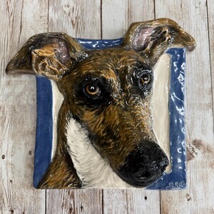 Può includere: Una scultura da parete in ceramica di una testa di cane. Il cane ha il pelo marrone e bianco e gli occhi marroni. La scultura è posizionata su uno sfondo blu.