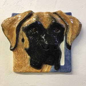 Può includere: Scultura in ceramica a rilievo raffigurante il volto di un cane, con una combinazione di colori marrone e nero. La testa del cane è centrata, con occhi dettagliati e una finitura lucida. La scultura è montata su una piastrella quadrata blu con un bordo bianco.