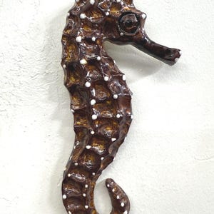 Op de afbeelding: Een bruin zeepaardbeeld met witte stippen. Het zeepaardje heeft een textuur en is op een witte achtergrond gemonteerd. Het beeld is een decoratief item, mogelijk voor een kust- of nautisch thema.