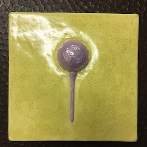 May include: Square ceramic tile with a light green background and a raised purple flower design. The flower has a spherical head and a long, slender stem. The tile has a speckled texture.
