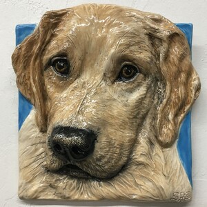 以下が含まれることがあります： ゴールデンレトリバーの犬の頭部のセラミック製の壁彫刻。犬は茶色の目と薄い茶色の鼻をしています。彫刻は青い背景に設置されています。