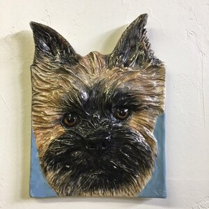 May include: A rectangular, textured ceramic sculpture of a dog's face. The dog has brown eyes, a black muzzle, and tan and black fur. The sculpture is set against a light blue background.