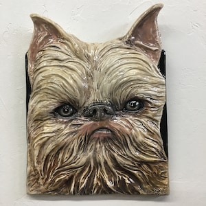 May include: A ceramic sculpture of a Brussels Griffon dog's face. The sculpture is painted in shades of brown, beige, and black, and has a realistic texture. The dog's eyes are open and looking directly at the viewer. The sculpture is signed "Sao" in the lower right corner.