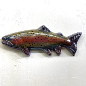 Può includere: Una scultura da parete in ceramica raffigurante una trota iridea con finitura lucida. Il pesce presenta un dorso verde oliva scuro, una marcata striscia laterale rosso-rosata e macchie nere sul corpo. Le pinne e la coda sono scure e violacee.