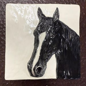 Könnte beinhalten: Eine schwarze und weiße Keramikfliese mit einem erhabenen Bild eines Pferdekopfes. Das Pferd hat einen weißen Streifen auf der Schnauze und eine schwarze Mähne und einen schwarzen Schweif.
