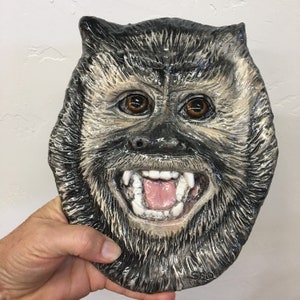 May include: A ceramic sculpture of a monkey face with a wide, toothy grin. The monkey has brown eyes, a black nose, and a gray and black fur texture. The sculpture is held in a hand.