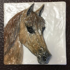 Könnte beinhalten: Eine Keramikfliese mit einer 3D-Reliefskulptur eines braunen Pferdekopfes. Das Pferd hat einen weißen Stern auf der Stirn und schaut nach links. Die Fliese ist weiß mit einer glänzenden Oberfläche.