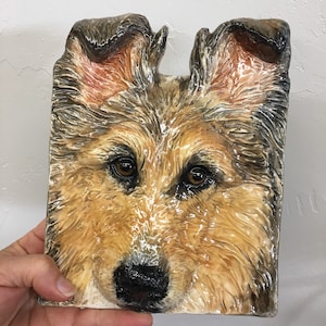 以下が含まれることがあります： 犬の顔のセラミック製の壁飾り。犬は茶色と白の毛並みで、長い鼻と大きく暗い目をしています。彫刻は細部までこだわっており、リアルな質感の毛並みと光沢のある仕上げが施されています。