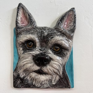 Può includere: Un'applique da parete in ceramica con un ritratto di un cane Schnauzer. Il cane ha un mantello nero e grigio, occhi marroni e un naso nero. Lo sfondo è un rettangolo azzurro, montato su un muro bianco.