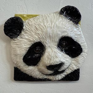 May include: A square ceramic panda face with black and white fur, black ears, and brown eyes. The panda's nose is black, and the background is a light colour. The panda has a yellow accent on the top.