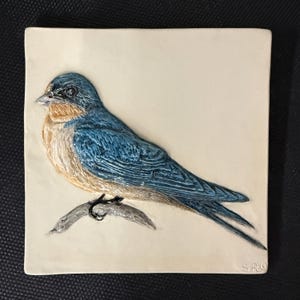 May include: A square, cream-coloured ceramic tile with a detailed relief of a blue and tan bird perched on a grey branch. The bird has intricate feather detailing against a plain background. The artist's initials are in the bottom right corner.