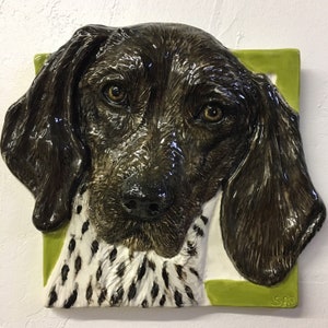 Könnte beinhalten: Eine Keramikskulptur eines Deutsch Kurzhaar Hundes mit braunem und schwarzem Fell und weißen Abzeichen. Der Kopf des Hundes ist nach vorne gerichtet, die Ohren hängen herunter. Die Skulptur ist vor einem grünen Hintergrund platziert.