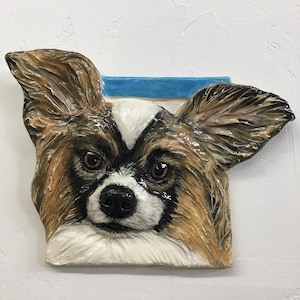 Può includere: Targa da parete in ceramica raffigurante il volto di un cane Papillon. Il cane ha pelo marrone e bianco, occhi grandi e un naso nero. Un rettangolo blu è sopra la testa del cane. Firma in basso a sinistra.