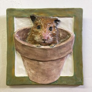 Può includere: Una piastrella da parete in ceramica con una cornice verde che raffigura un criceto marrone che sporge da un vaso di terracotta.