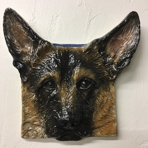 以下が含まれることがあります： ジャーマンシェパード犬の顔を描いた正方形の壁掛け。犬の毛皮は茶色、黒、タンの色合いで表現され、大きな直立した耳があります。犬の目は暗く表現力豊かで、鼻は黒です。
