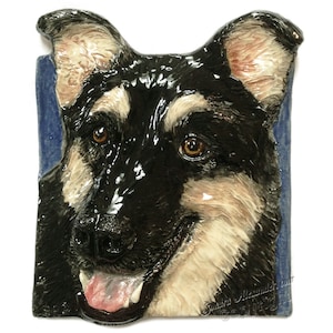 Op de afbeelding: Een keramisch reliëfsculptuur van de kop van een Duitse herdershond. De hond heeft zwart en bruin haar, bruine ogen en een roze tong. De sculptuur is geplaatst tegen een blauwe achtergrond en gesigneerd "Sonora Alexander" met een copyrightvermelding.