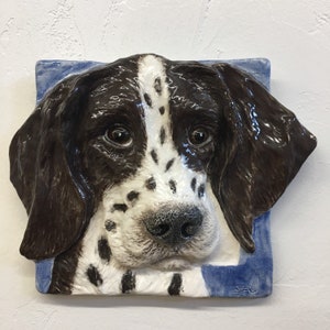 Può includere: Una scultura da parete in ceramica di un cane marrone e bianco con macchie nere. La testa del cane è inclinata di lato e i suoi occhi sono chiusi. La scultura è posizionata su uno sfondo blu.
