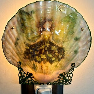 May include: A wall plug-in night light featuring a large, iridescent seashell shade. The shell is decorated with a printed image of a fairy with green wings and a dark floral dress. It is held by a dark green metal bracket and emits a warm glow.