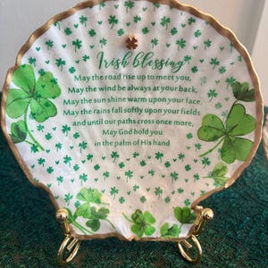 May include: A decorative, shell-shaped plaque with a gold trim and stand. The white surface is adorned with green shamrocks and the text "Irish blessing". The plaque is a St. Patrick's Day themed item.