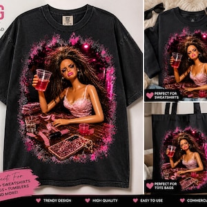 May include: A black t-shirt featuring a graphic print of a doll with messy brown hair wearing a pink lace top, holding a red drink. The graphic includes a glittery pink phone case, a lipstick, and small cups on a table, surrounded by a pink aura.