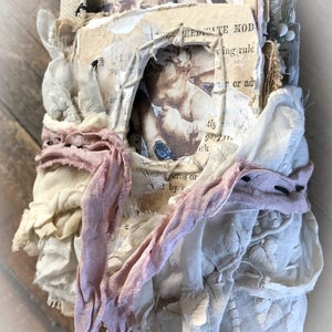 May include: A handmade journal with a rustic aesthetic. The journal is covered in layers of fabric, lace, and paper, and has a hole cut out of the front cover. The journal is tied together with a pink ribbon.