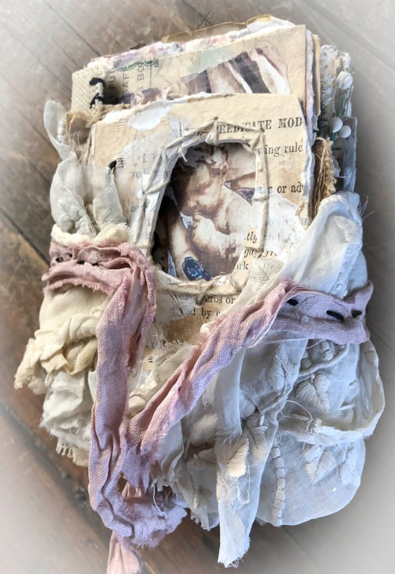 May include: A handmade journal with a rustic aesthetic. The journal is covered in layers of fabric, lace, and paper, and has a hole cut out of the front cover. The journal is tied together with a pink ribbon.