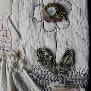 May include: A white fabric with a stitched flower design. The flower has a button in the center and is surrounded by fabric scraps. The fabric is stitched to a striped fabric with the word "grow" stitched on a tag.